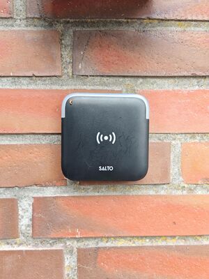 Bild eines schwarzen eckigen Wandlesers mit abgerundeten Ecken an einer Backsteinwand. Auf dem Wandleser ist die Aufschrift "Salto" sowie einem Funksymbol. An der oberen Seite befindet sich ein schmaler transparenter Kunststoffstreifen.